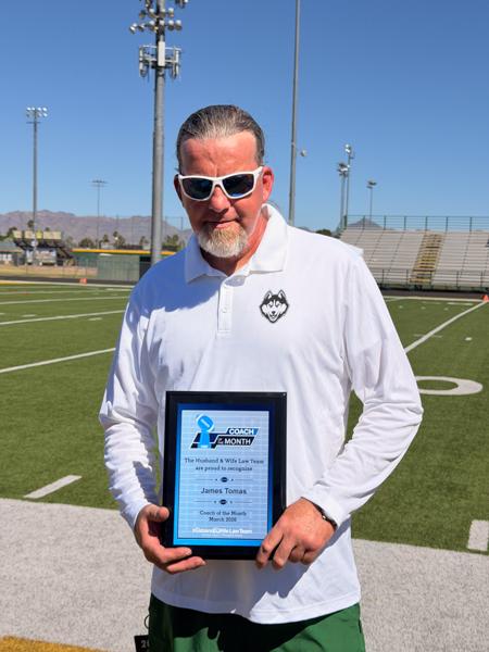 On a bright day at the football field, Coach Tomas poses with his “Coach of the Month” plaque, mountains and bleachers in the background.
