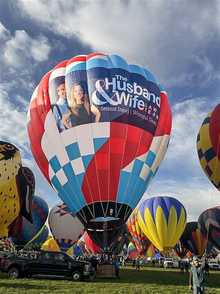 The vibrant red, white, and blue Husband & Wife Law Team balloon, featuring Mark and Alexis Breyer, joins a colorful lineup of balloons at the Albuquerque International Balloon Fiesta.