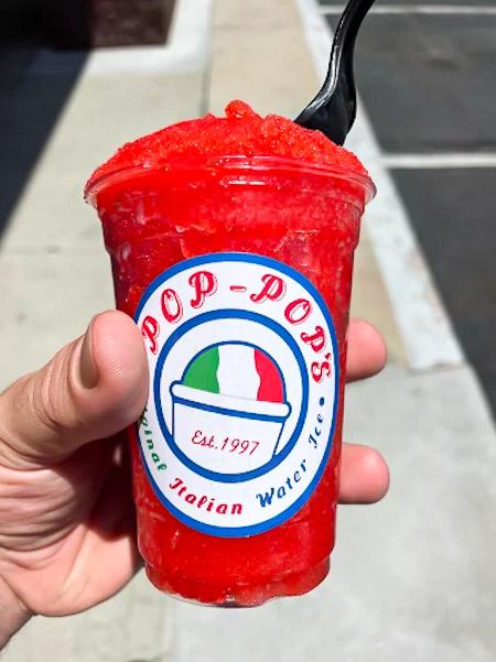 Hand holding a cup of red Italian ice with a black spoon, labeled "Pop-Pop's," featuring an Italian flag design.