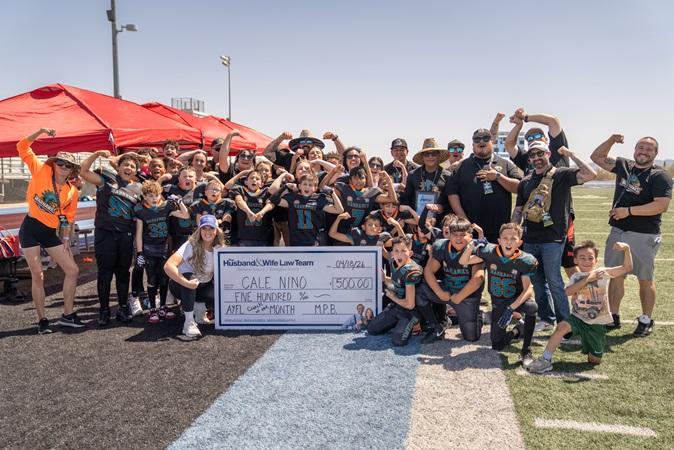 On the football field, AYFL athletes and supporters cheer for Coach Nino as he’s presented with a large check from the Husband & Wife Law Team.