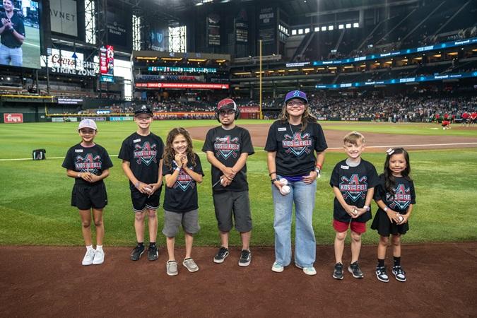 Group of children and an adult in matching shirts stand on a baseball field near the pitcher's mound at a stadium event.