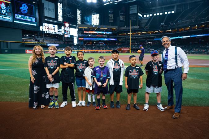 A group of children and two adults stand on a baseball field, smiling. The stadium is well-lit, with visible seating and signage in the background.