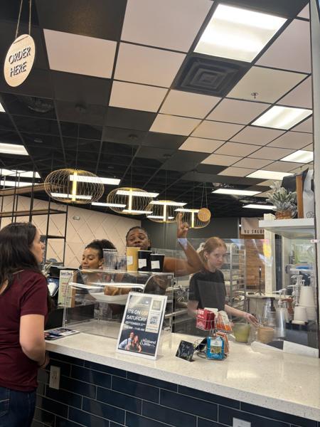 Customers order at a cafe counter with staff preparing drinks. Overhead lights and a "Order Here" sign are visible.
