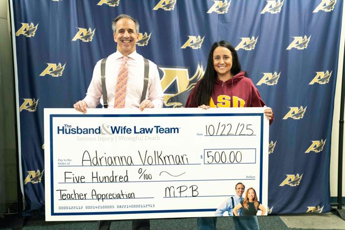 Mark Breyer and the Teacher of the Month hold a large check for $500 in front of a backdrop with logos. The check is for teacher appreciation.