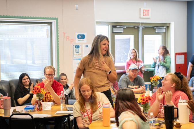 Sunrise Elementary faculty sit at tables in a bright room. Ms. Shade stands, smiling with surprise look and her hand on her chest, as others look on and smile.